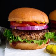 Turkey Burger layered with bun, ketchup, lettuce, turkey patty, red onion, tomato and yellow mustard. Served with a side of pickles on a white platter.