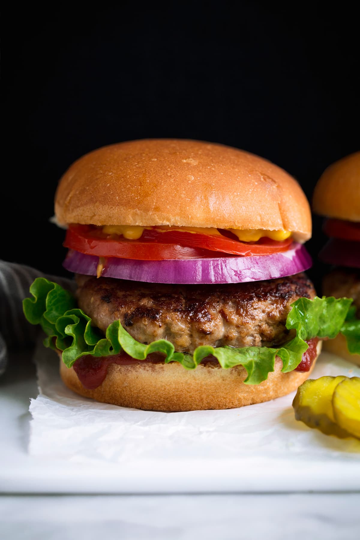 Turkey Burger layered with bun, ketchup, lettuce, turkey patty, red onion, tomato and yellow mustard. Served with a side of pickles on a white platter.