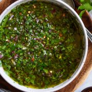 Overhead close up image of chimichurri in a bowl set over a wooden plate on a marble surface.