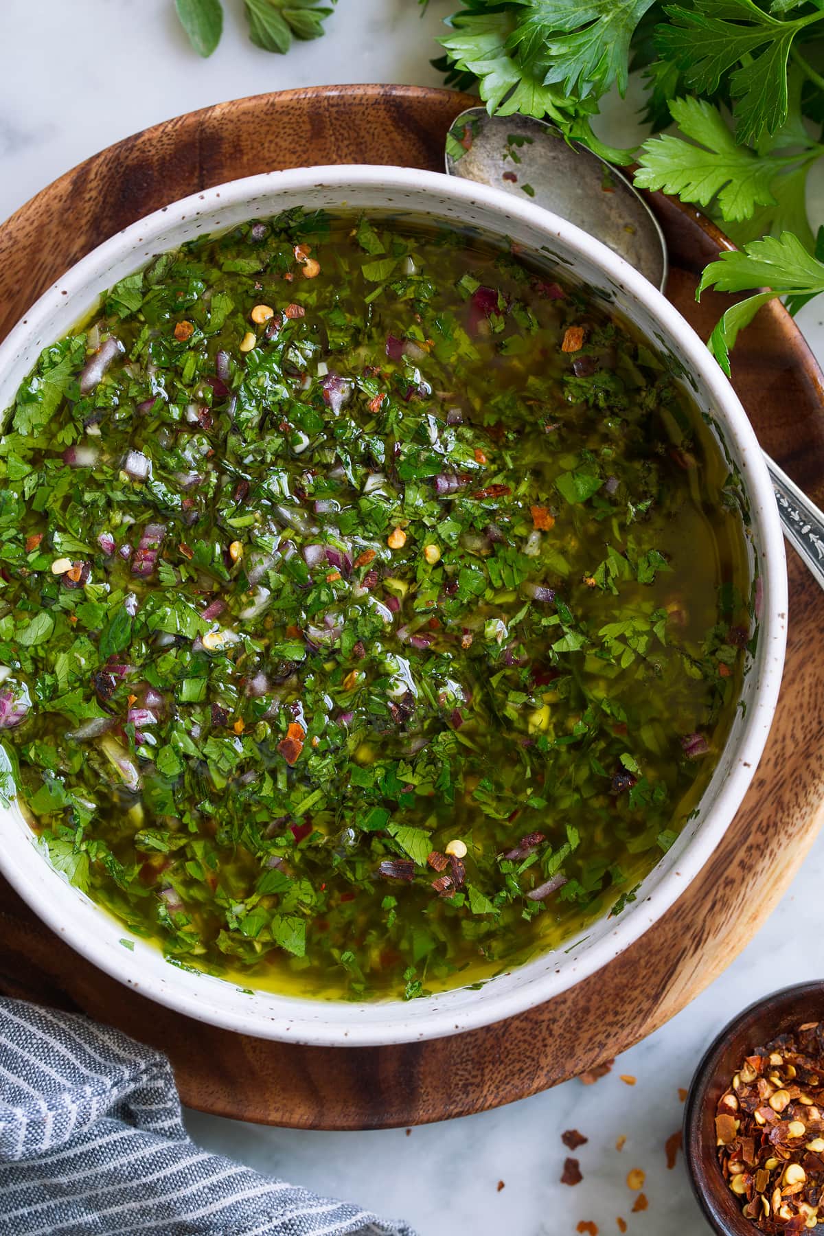 Overhead close up image of chimichurri in a bowl set over a wooden plate on a marble surface.