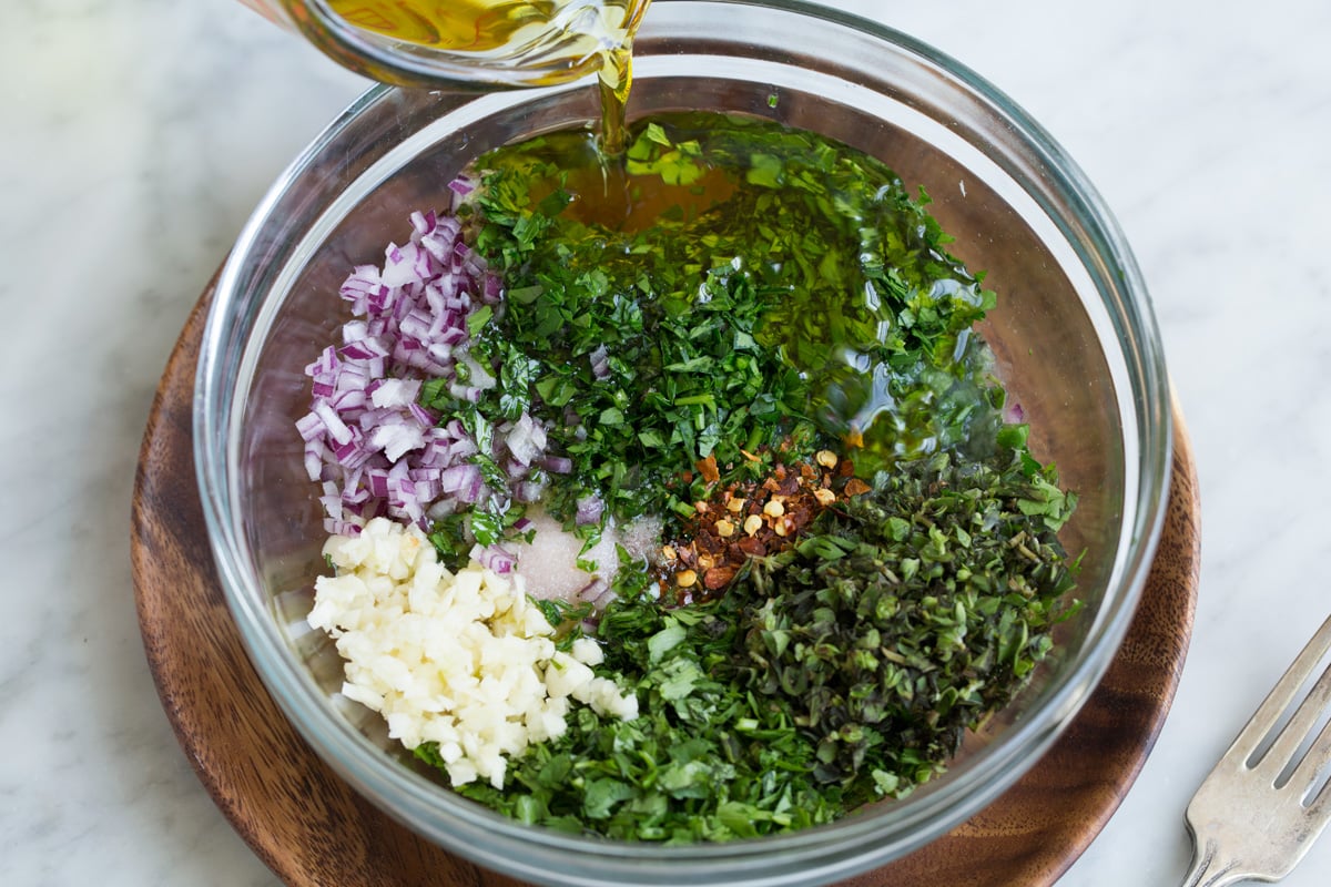Showing how to make chimichurri. Mixing parsley, cilantro, oregano, onion, garlic, red pepper flakes, olive oil, lemon and red wine vinegar in a glass mixing bowl. 