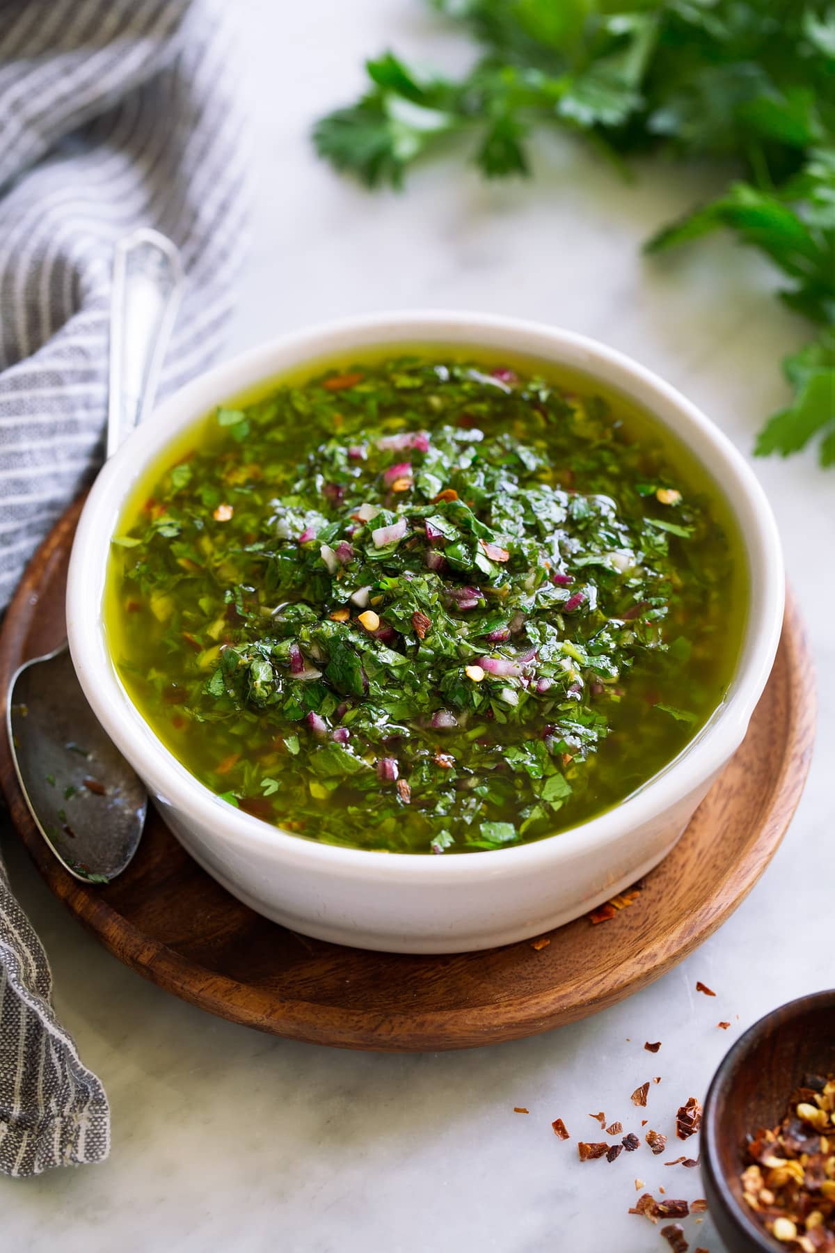 Chimichurri sauce in a small white bowl set over a wooden plate with a spoon to the side.