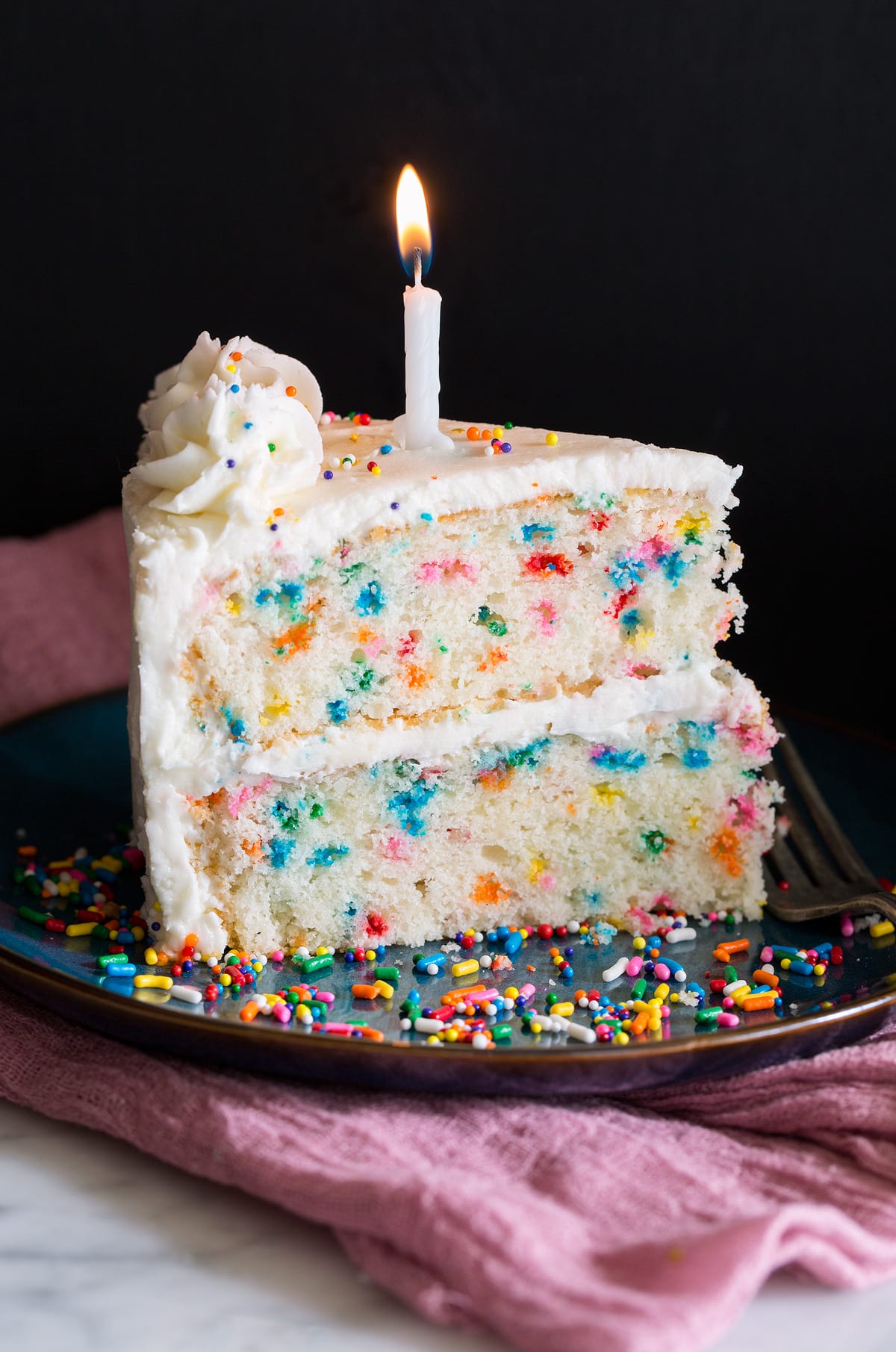 Slice of birthday cake with multi color dots in cake and topped with buttercream frosting and a birthday. candle.