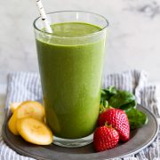 Green smoothie in a tall glass with strawberries, spinach and bananas to the side.