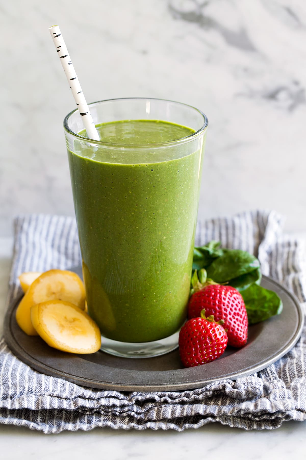 Green smoothie in a tall glass with strawberries, spinach and bananas to the side.