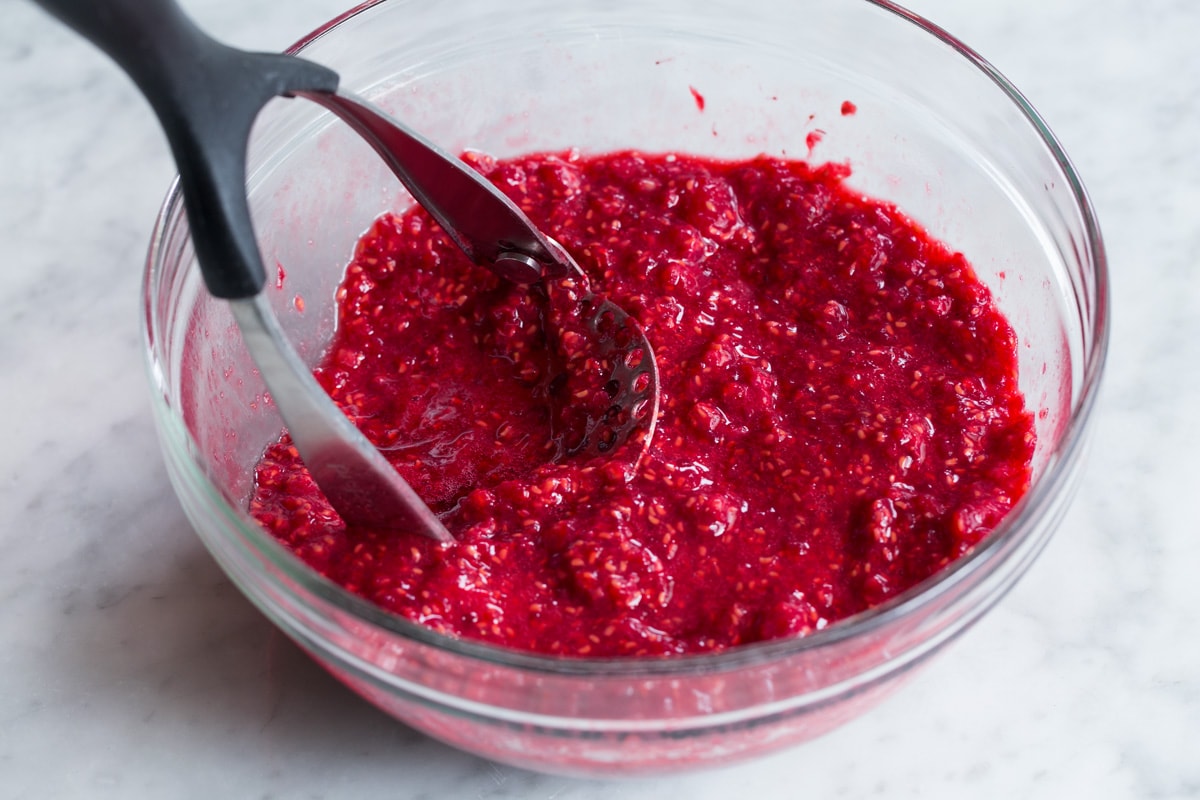 Showing how to make raspberry jam. Staring here by crushing raspberries in a glass mixing bowl with a potato masher.