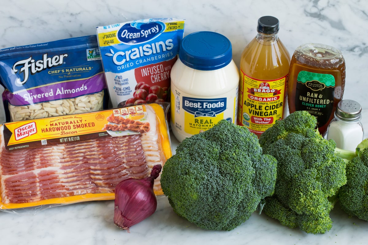 Ingredients to make broccoli salad shown here including broccoli, salt, honey, apple cider vinegar, mayonnaise, dried cranberries, slivered almonds, bacon and red onion.