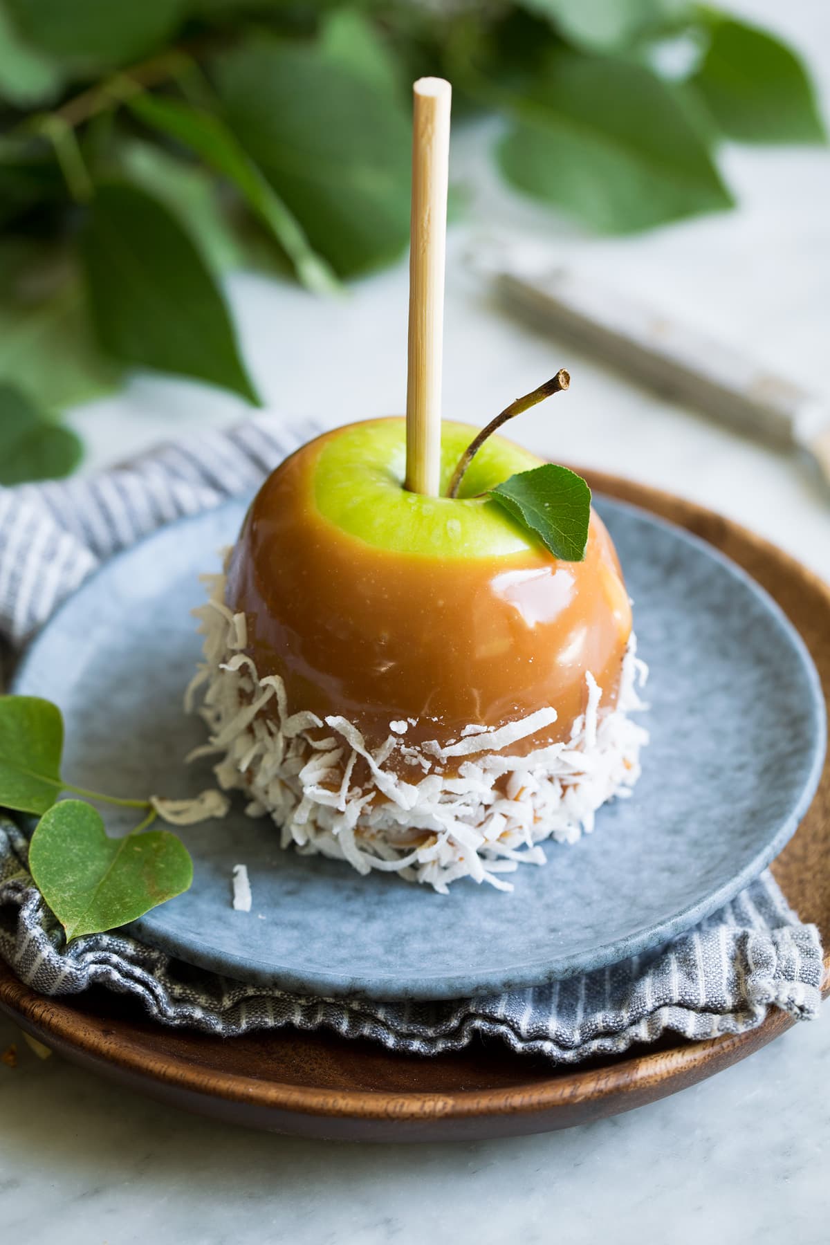 Single caramel apple rolled in shredded coconut sitting on a blue plate on top of a wooden plate.