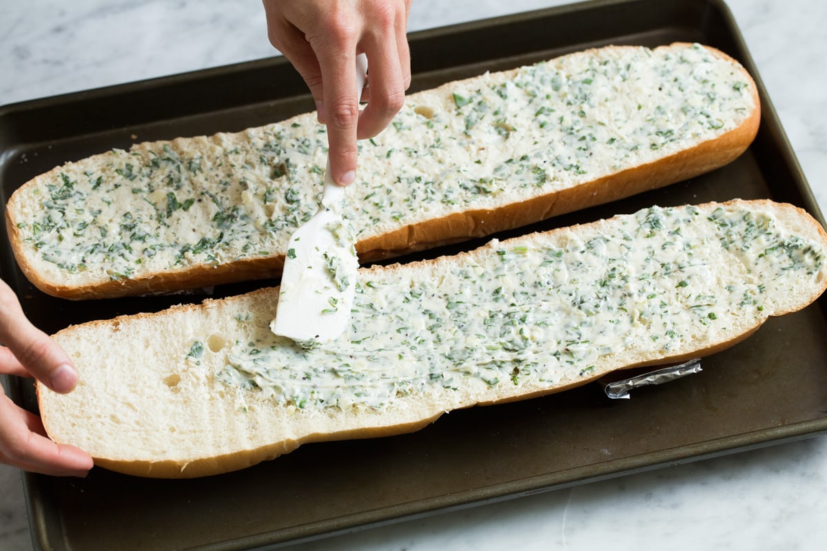 Garlic Bread - super fast, super easy and incredibly flavorful! Skip the margarine coated garlic bread from the store and make this butter version instead, it's a hundred times better! #garlicbread #sidedish #bread #appetizer Spreading garlic butter over french bread halves on a baking sheet.