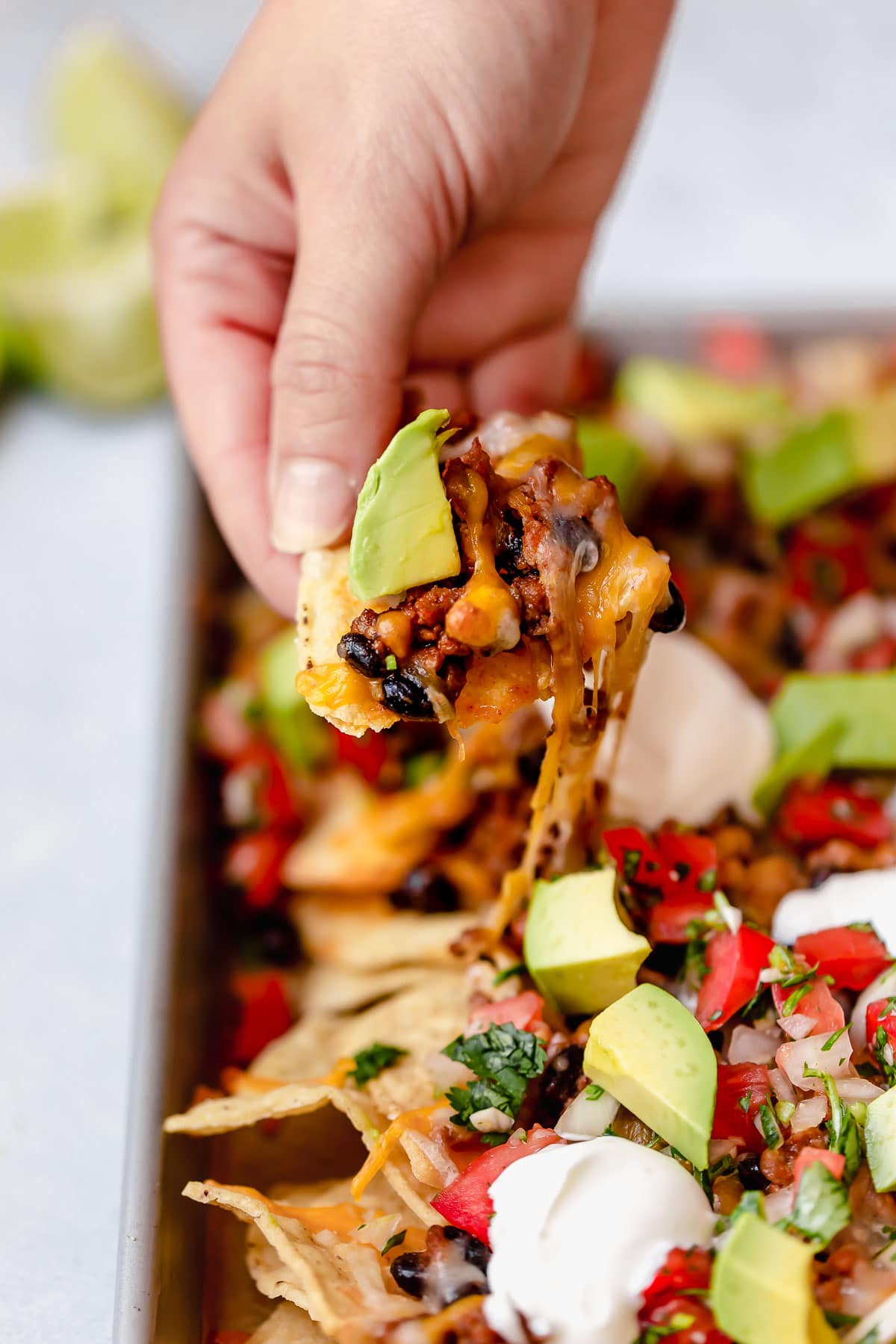 Easy Nachos - layered with seasoned beef and black beans, melty cheese, avocado and sour cream. SO GOOD! #nachos #sheetpan #beef #blackbeans #dinner #recipe Hand holding nacho chip with melted cheese.