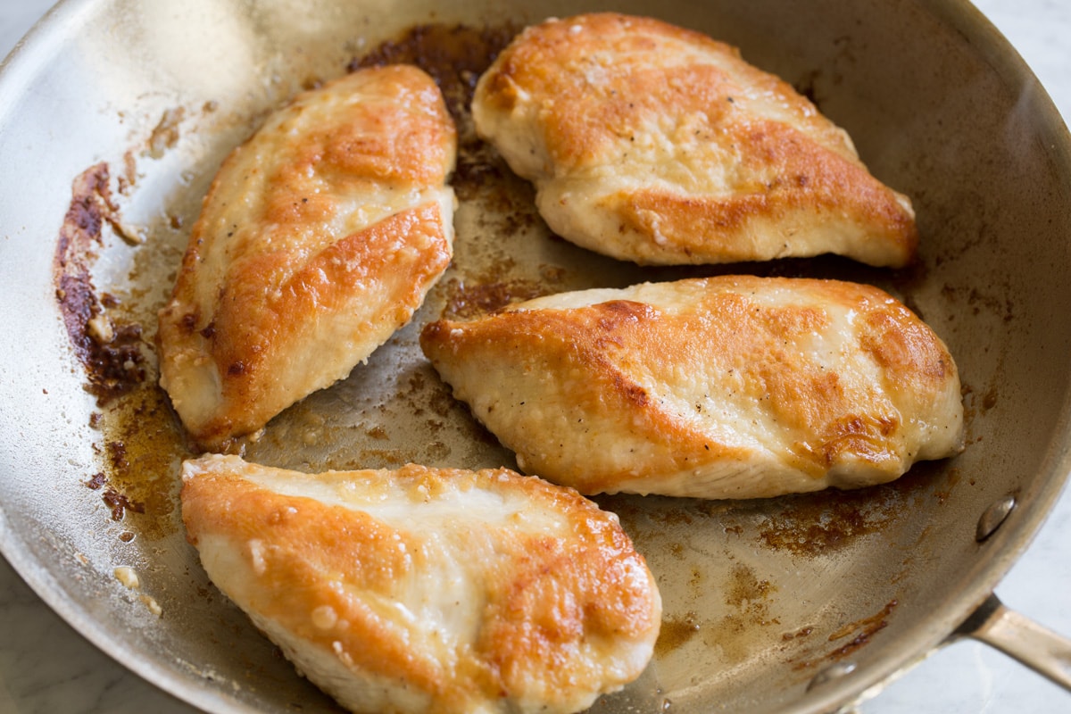 Four golden brown seared chicken breasts in a large stainless steel skillet.