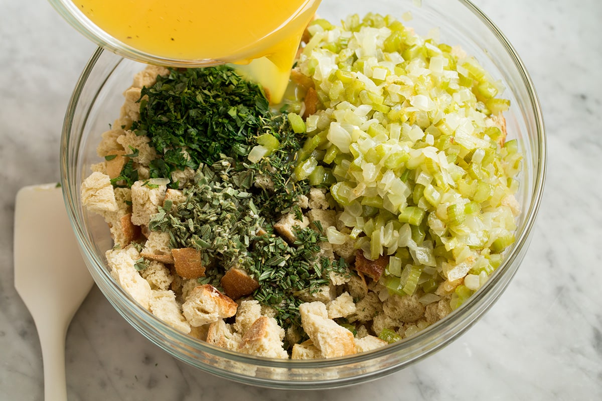 Showing how to make stuffing. Pouring broth mixture over dried bread cubes, celery mixture and herbs in a glass mixing bowl.