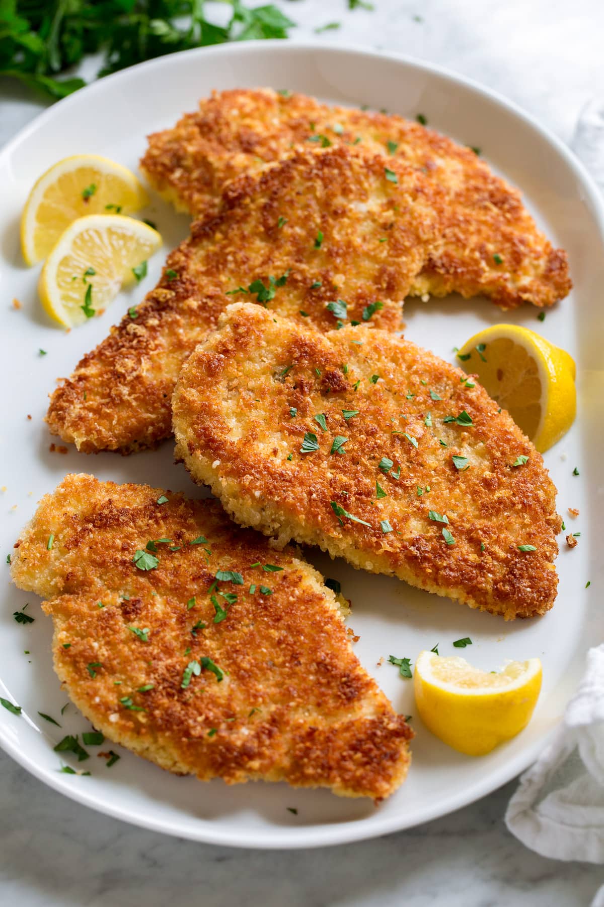 Parmesan Crusted Chicken cutlets shown on a white serving platter from a side angle. Lemons are shown to the side and parsley is garnished over.