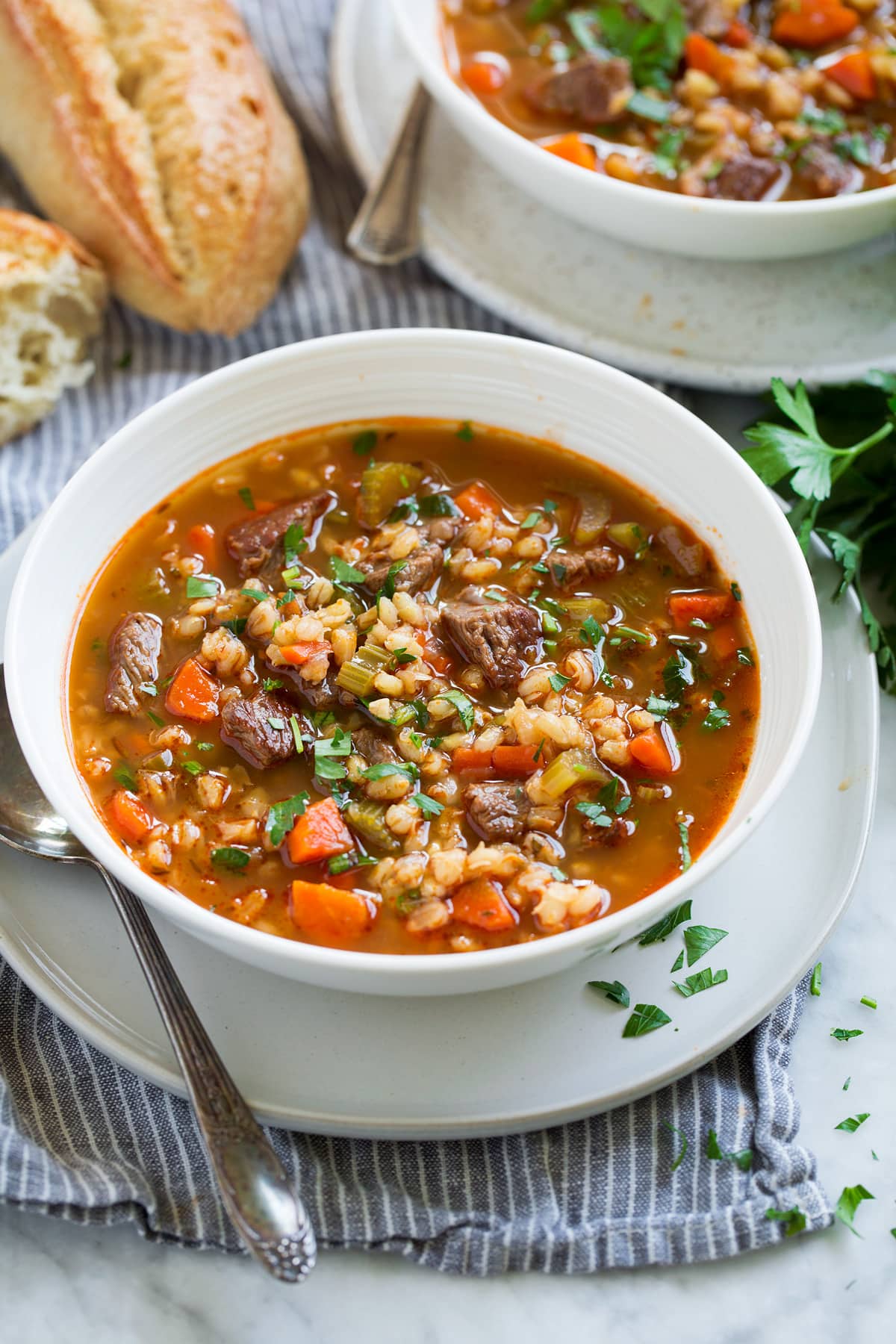 White bowl set over a white plate filled with beef and barley soup with a second serving shown in the background. Also shown is a side serving suggestion of fresh bread.