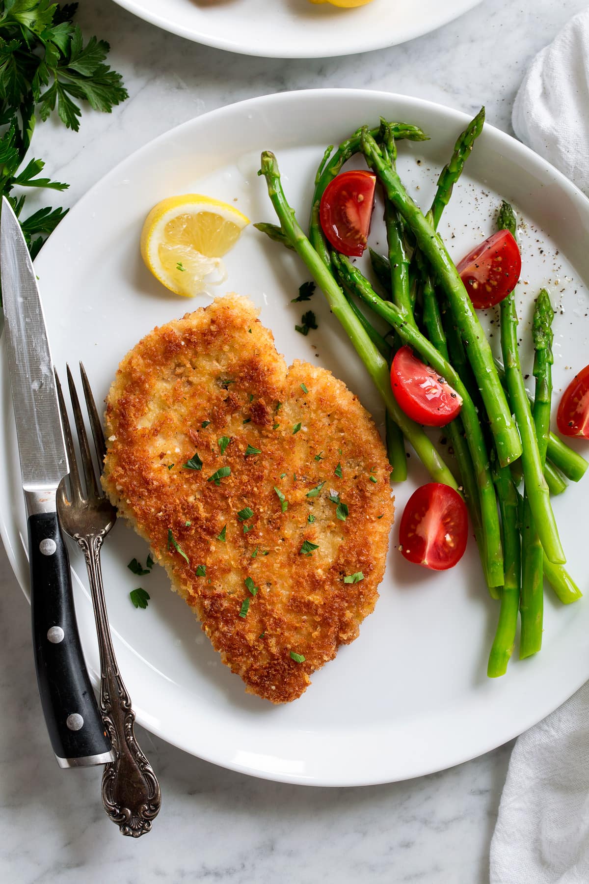 Parmesan Crusted Chicken fried breast shown on a serving plate with a side of asparagus and tomatoes.