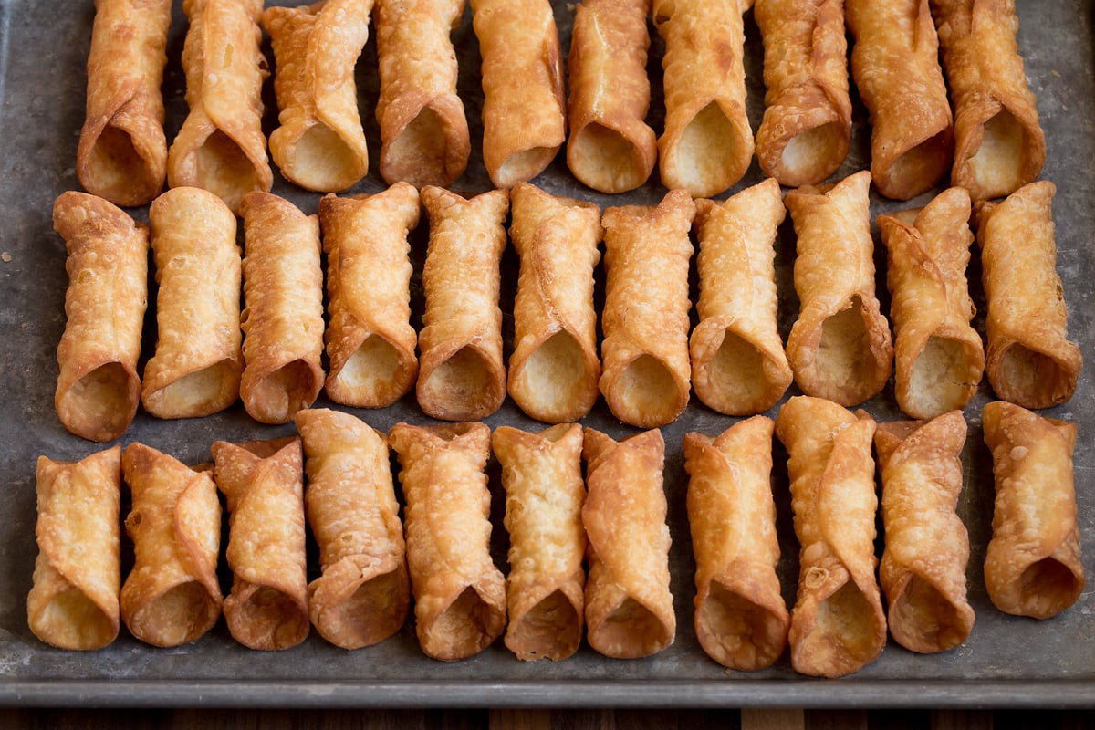 Homemade fried cannoli shells on a baking sheet shown before filling.