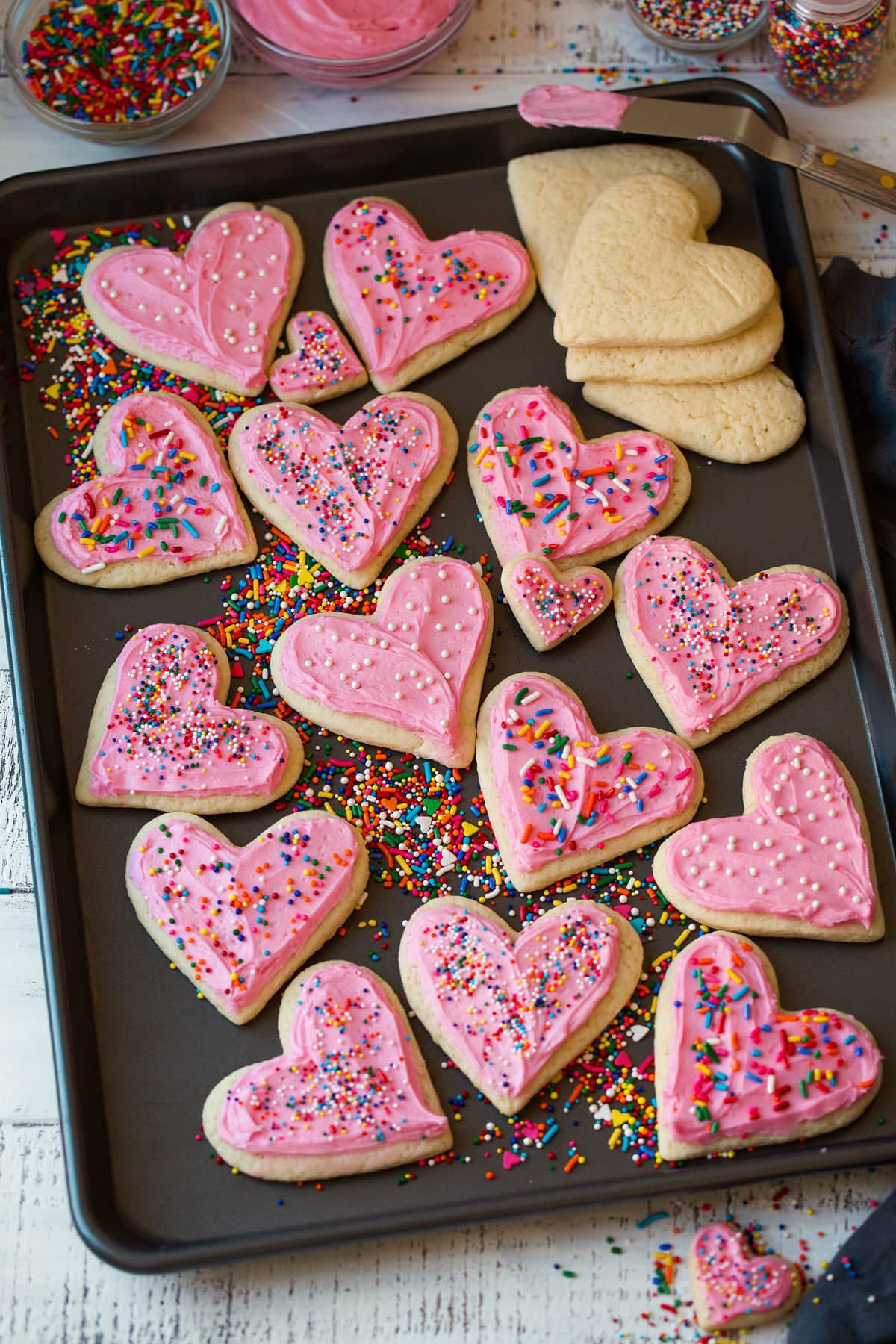 Cutout sugar cookies in heart shapes on a baking sheet shown frosted with sprinkles.