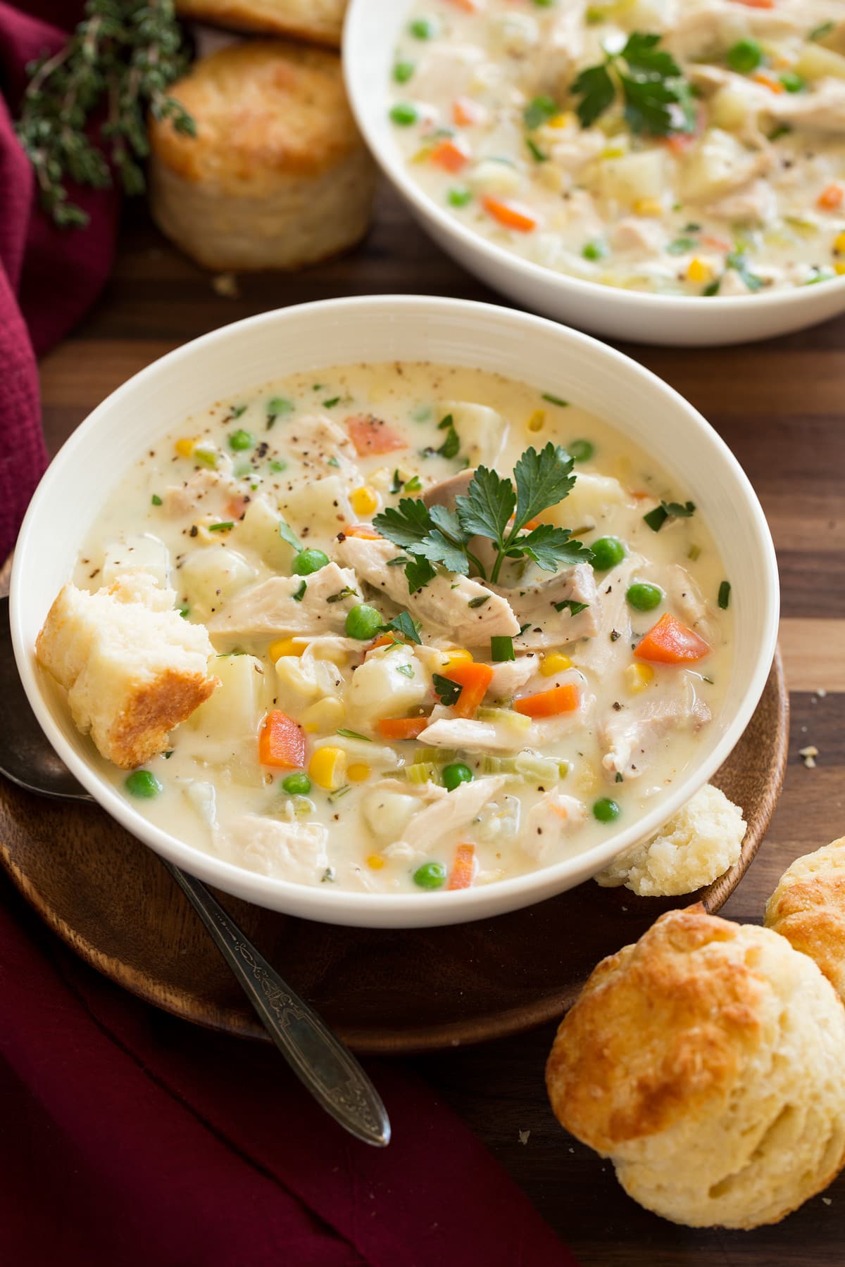 Two bowls of chicken pot pie soup in white serving dishes set over wooden a wooden tabletop. Biscuits are served to the side.