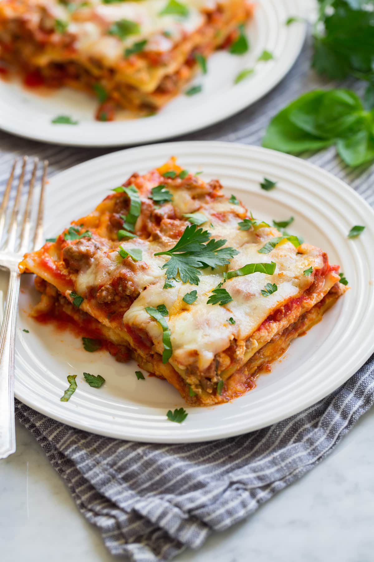 Slice of easy lasagna on a white serving plate with a second serving shown in the background. Plates are sitting on a grey striped napkin on a marble surface.