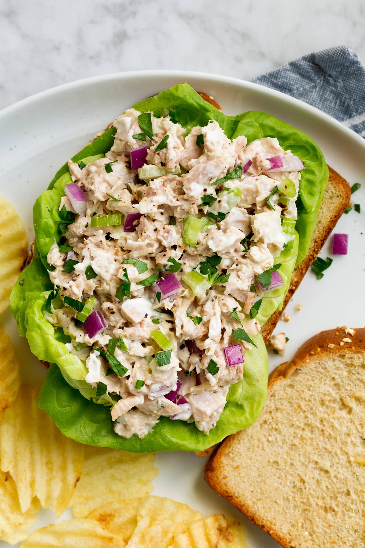 Tuna salad atop bread slices and lettuce leaves. Shown on a white plate with a side of potato chips.