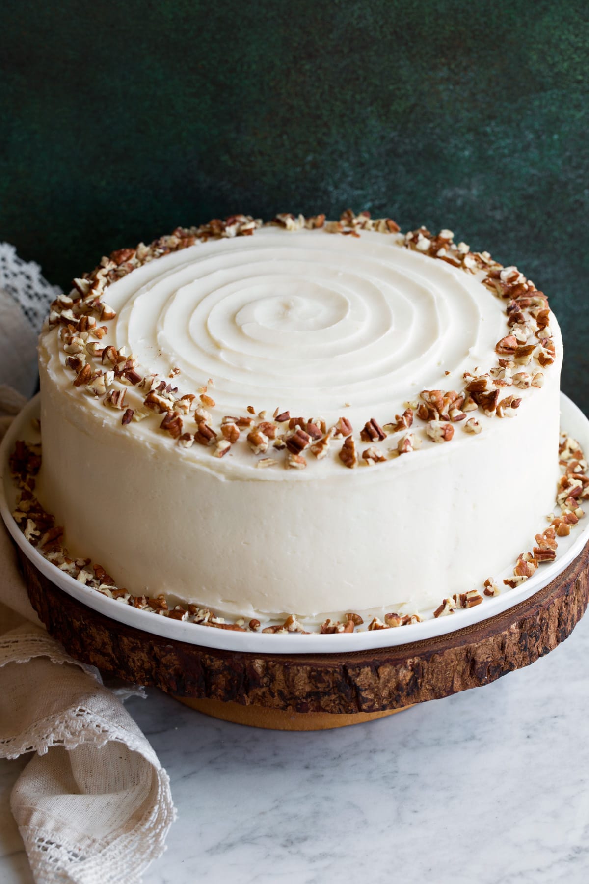 Cake on a wooden cake stand covered in cream cheese frosting and garnished with a wring of chopped pecans on top.