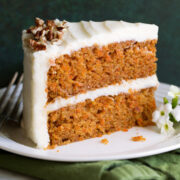 Slice of carrot cake with cream cheese frosting on a white dessert plate sitting on a green cloth napkin.