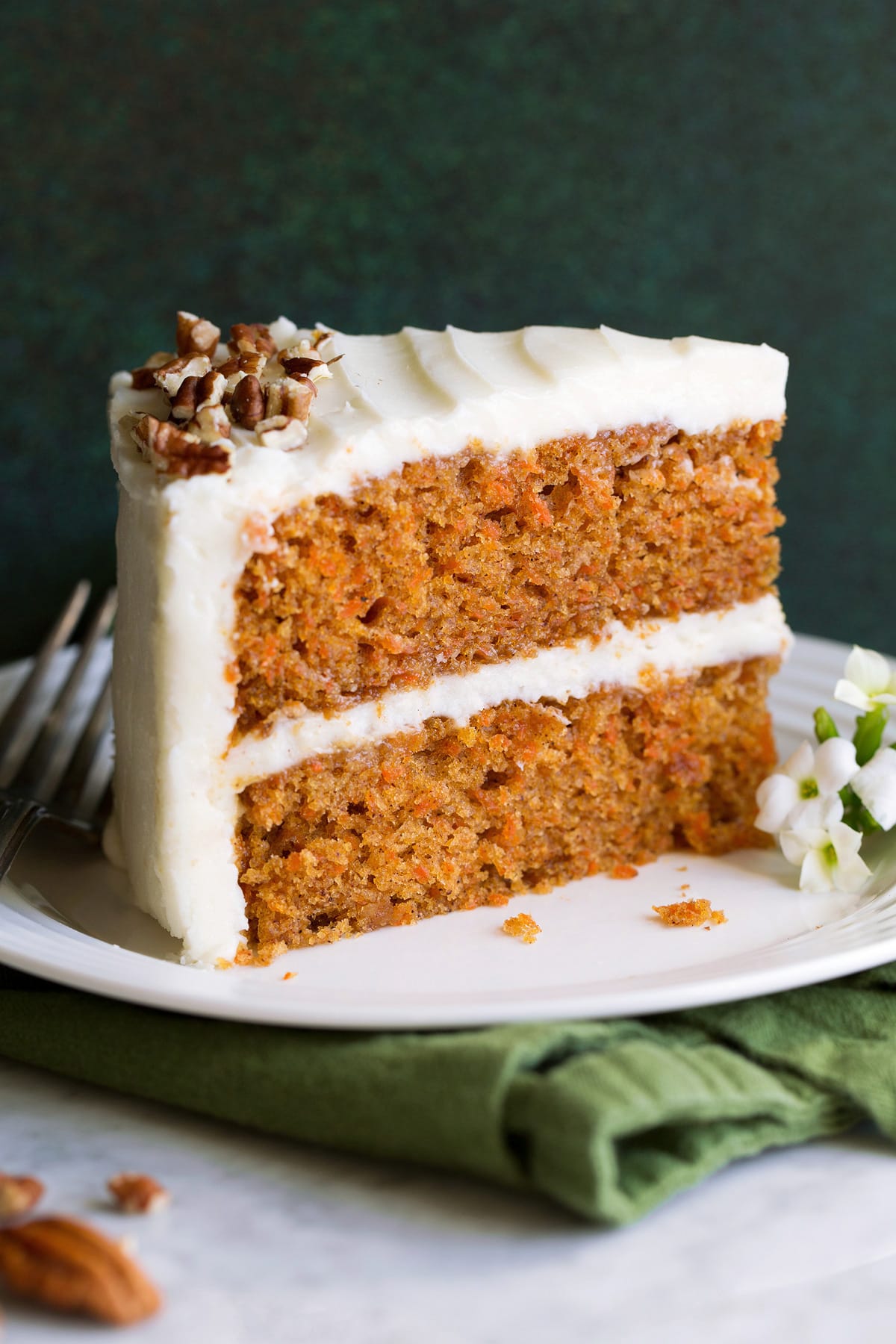 Slice of carrot cake with cream cheese frosting on a white dessert plate sitting on a green cloth napkin.