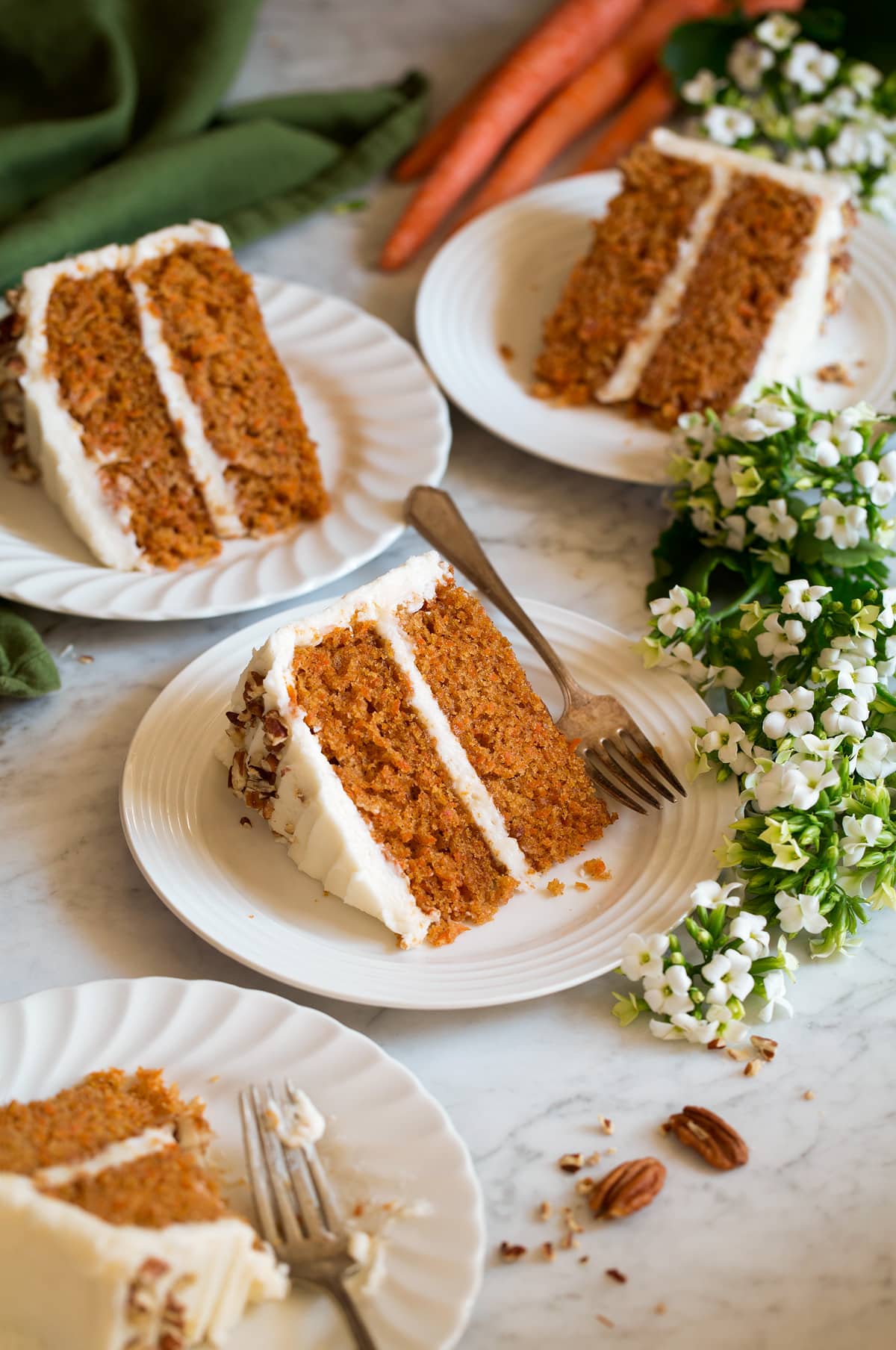 Best Ever Carrot Cake Four slices of carrot cake shown on white scalloped dessert plates with flowers and carrots around plates.