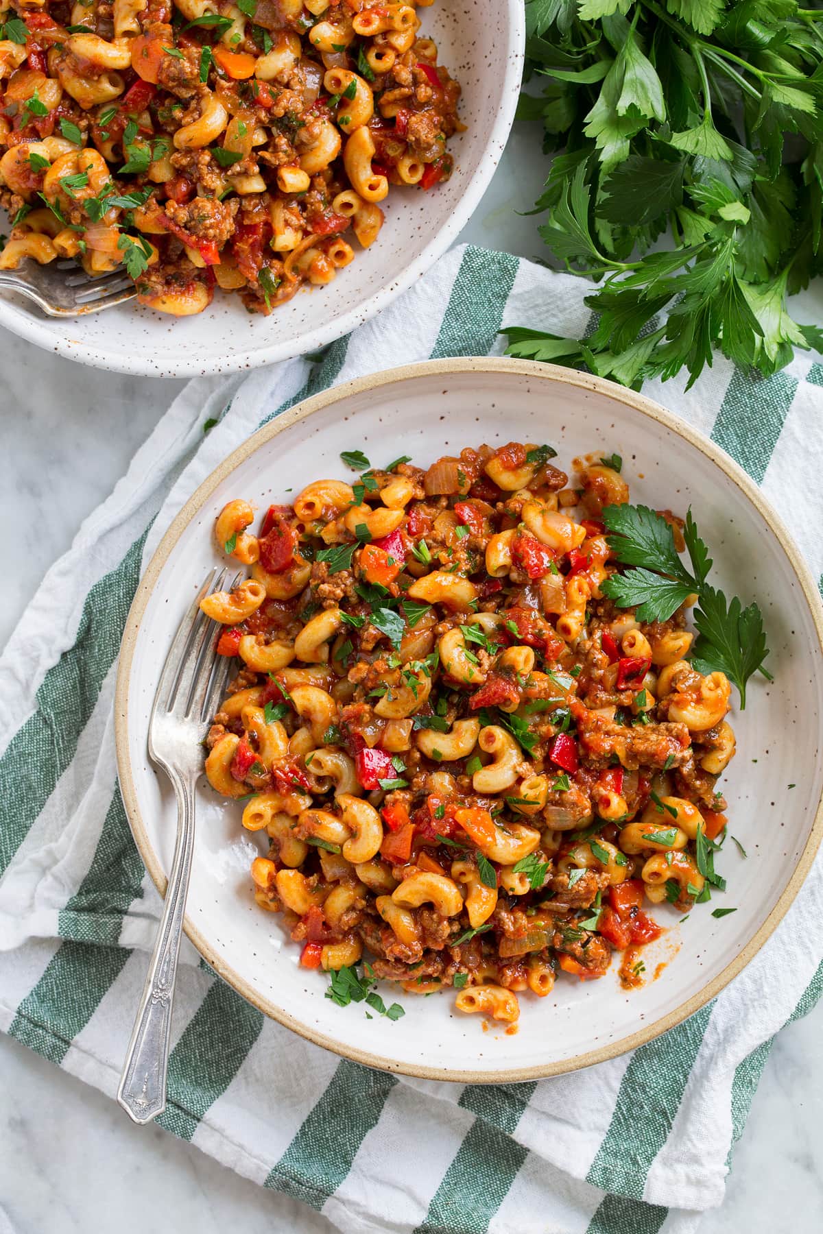 Serving of ground beef and macaroni goulash shown overhead in a white pasta bowl set over a green and white stripped napkin with parsley to the side.