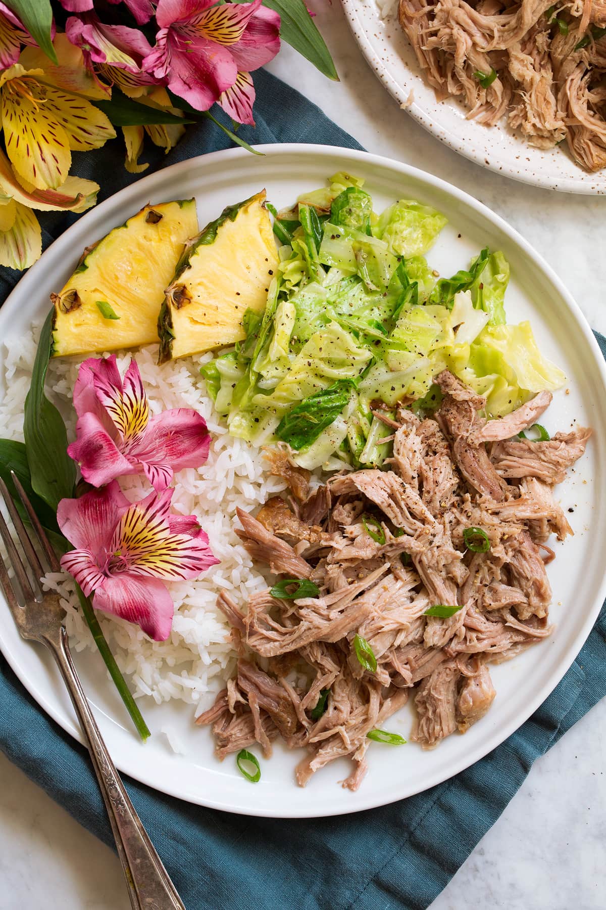 Single serving of shredded kalua pork on a white serving plate shown with sides of white rice, sautéed cabbage, fresh pineapple and flowers.