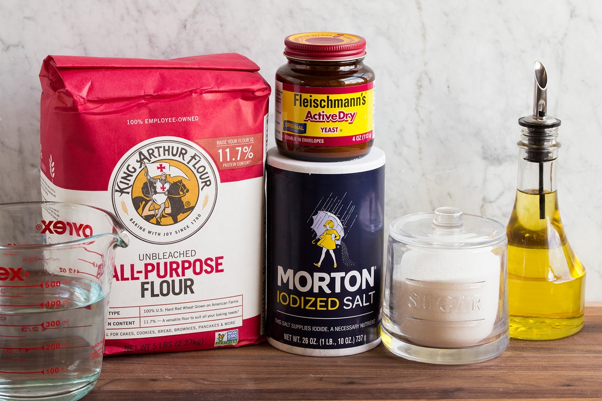 Image of ingredients used to make pizza dough. Includes all-purpose flour, water, salt, yeast, sugar and olive oil.