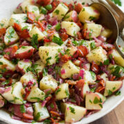German potato salad shown in a white bowl. A silver serving spoon and fork are resting to the side, bowl is sitting on a wooden board with parsley in the background.