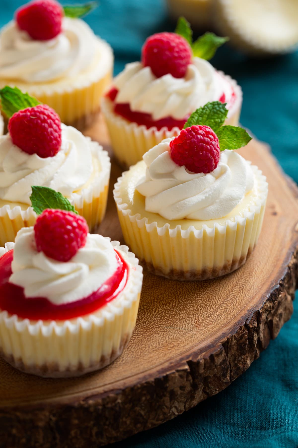 5 mini cheesecakes in cupcake liners sitting on a wooden serving platter set over a blue cloth.