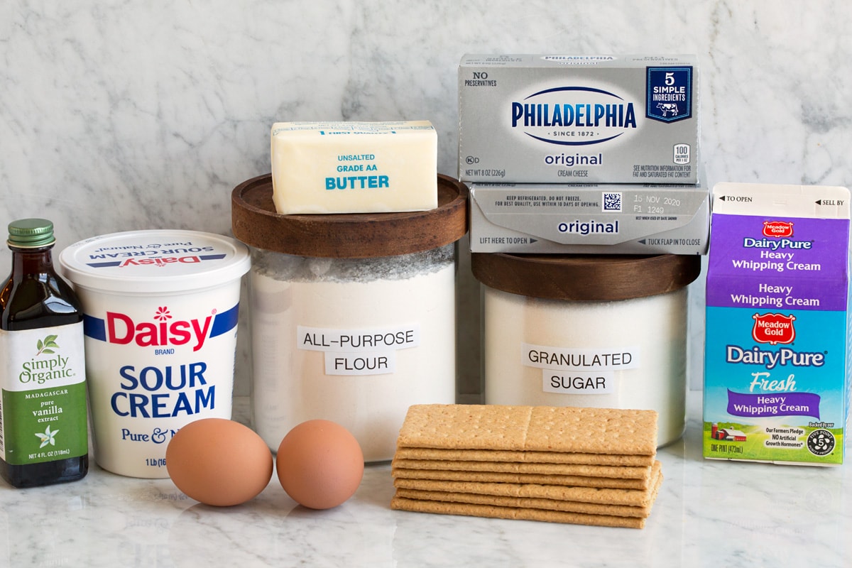 Image of ingredients used to make mini cheesecakes. Includes cream cheese, granulated sugar, all-purpose flour, eggs, graham crackers, sour cream, heavy cream, vanilla and butter.