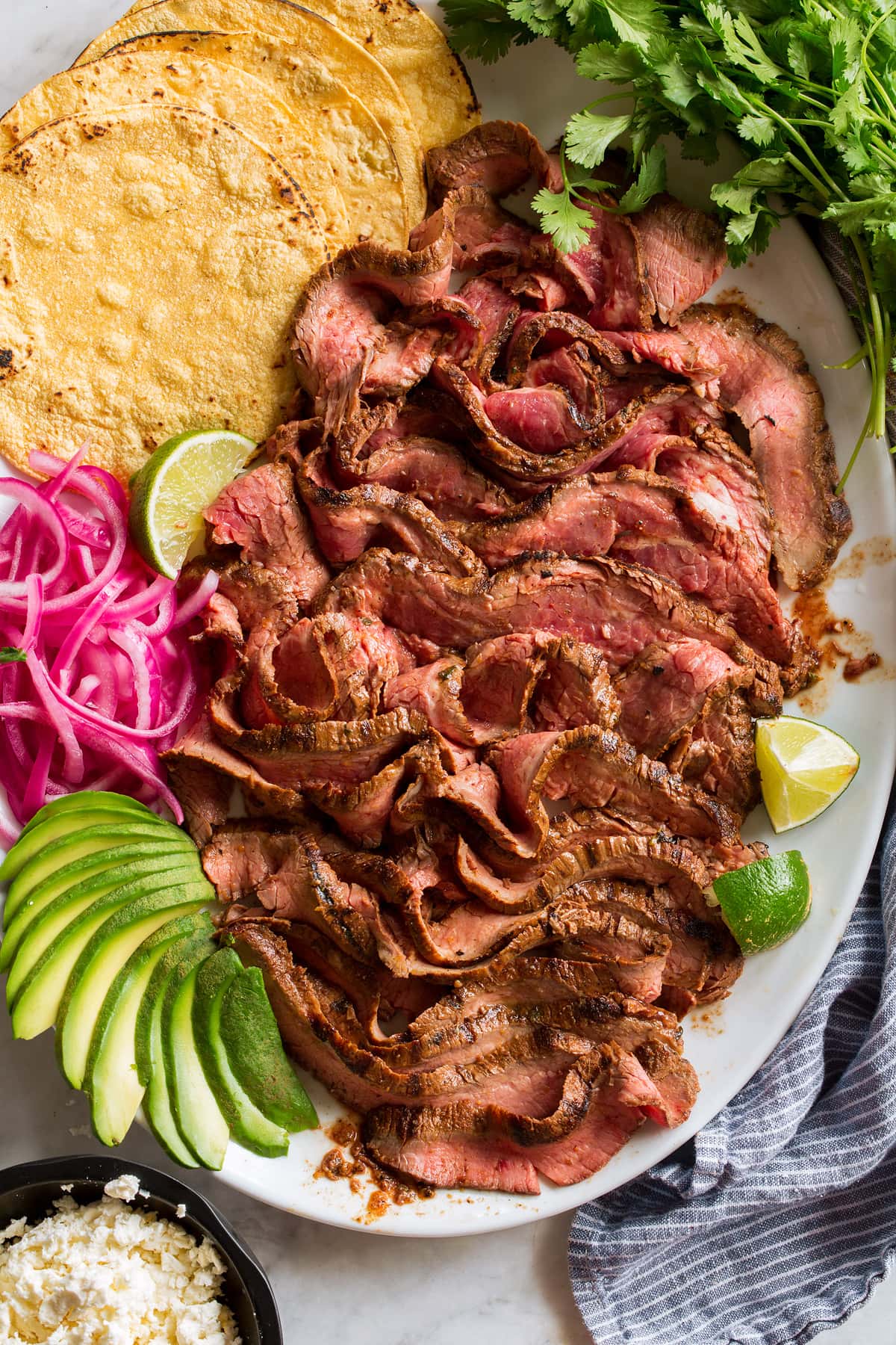Carne Asada Overhead image of thin sliced carne asada on a serving platter with tortillas, onions, avocado, lime and cilantro.