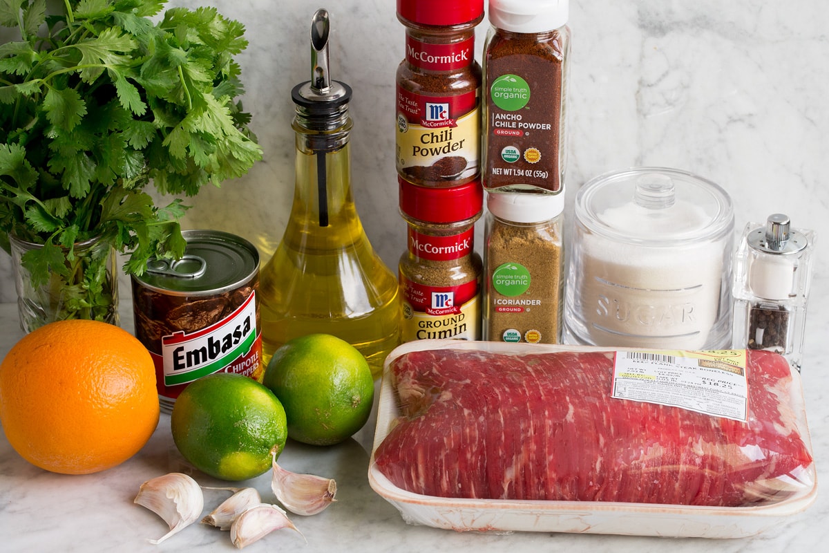 Carne Asada Image of ingredients used to make carne asada and marinade. Includes flank steak, cilantro, olive oil, chili powders, cumin, chipotle peppers, limes, oranges, sugar, garlic, salt and pepper.