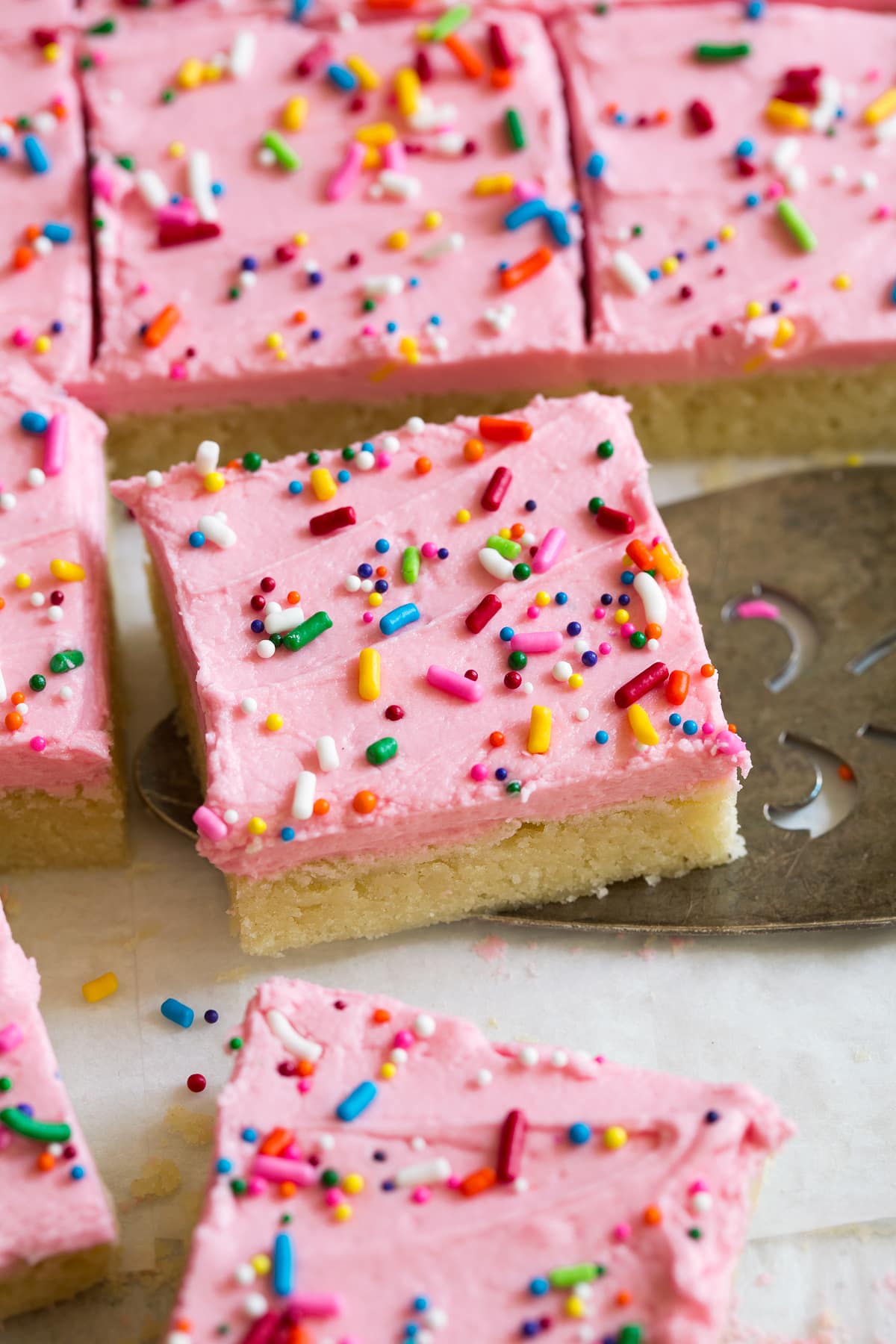 Close up image of sugar cookie bar decorated with pink frosting and sprinkles. It is being scooped up with a serving spatula.