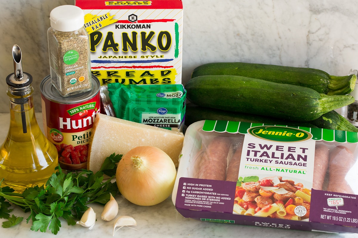 Image of ingredients used to make zucchini boats. Shows sausage, zucchini, onion, parmesan, parsley, garlic, mozzarella, tomatoes, Italian seasoning, panko bread crunbs and olive oil.