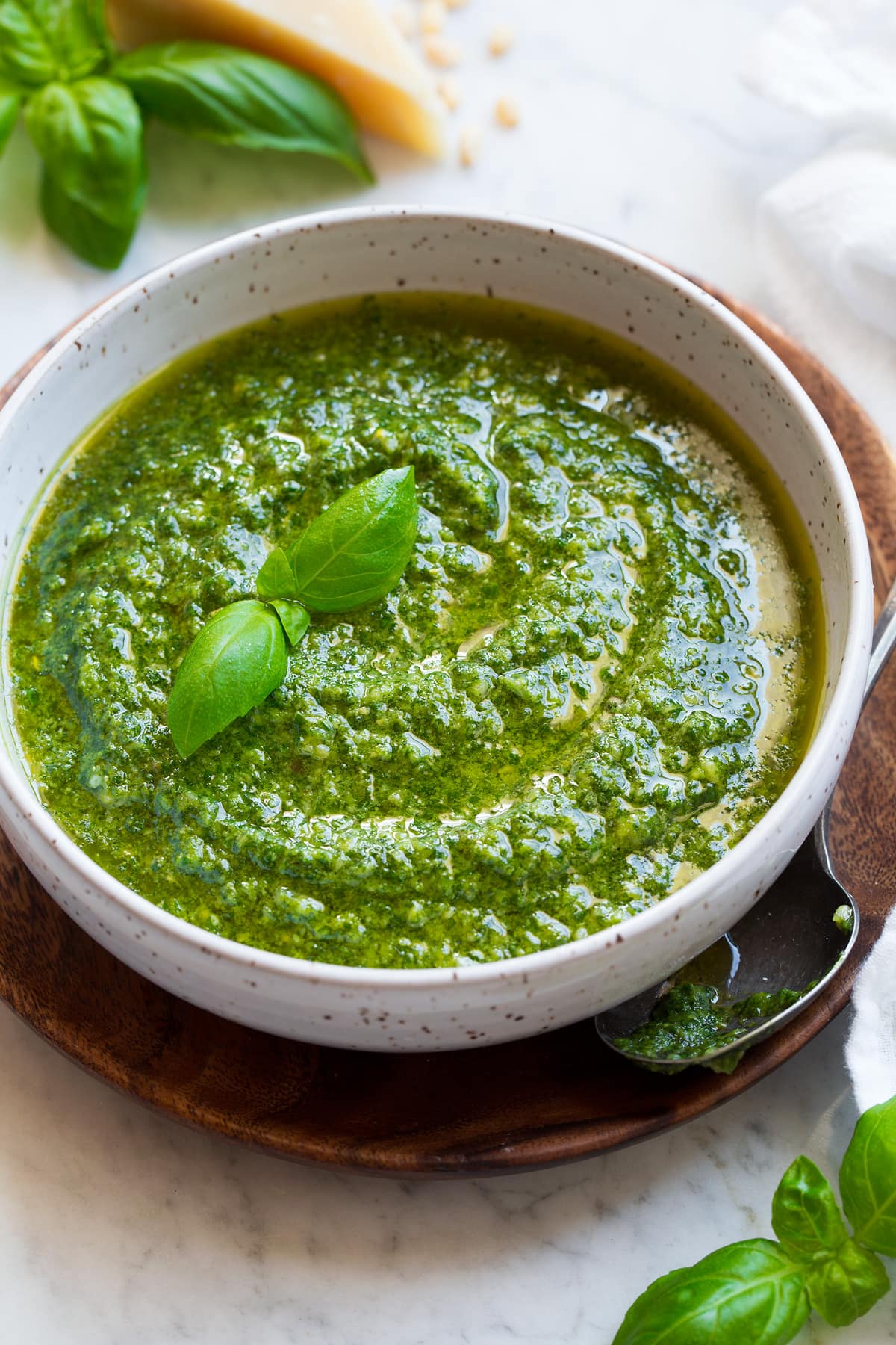 Bowl full of fresh homemade pesto. Bowl is resting on a wooden platter. Parmesan, fresh basil and pine nuts are shown in the background.