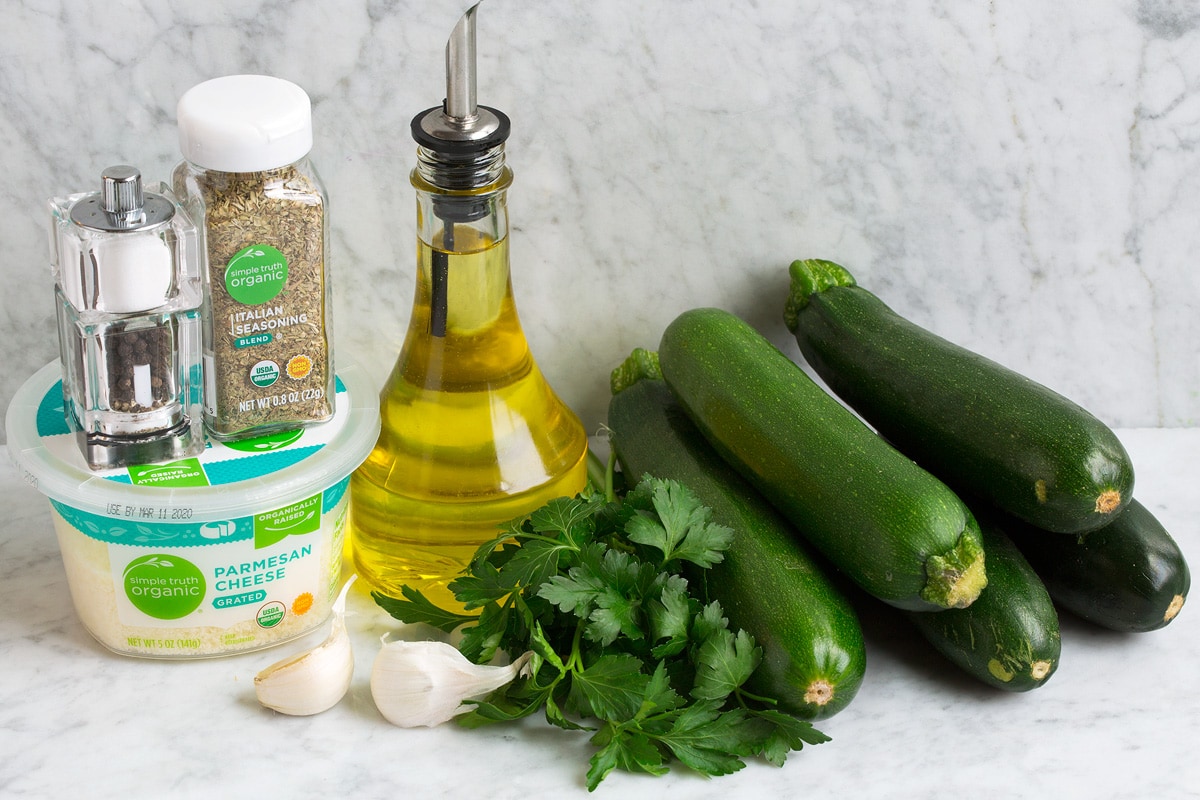 Image of ingredients used to make baked zucchini. Includes 5 medium zucchini, parsley, olive oil, Italian seasoning, salt and pepper, parmesan and garlic.
