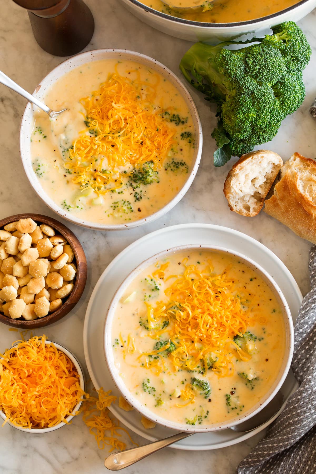 Image overhead of two servings of broccoli cheese soup in serving bowls. Crackers, broccoli, bread and cheese are shown to the side.