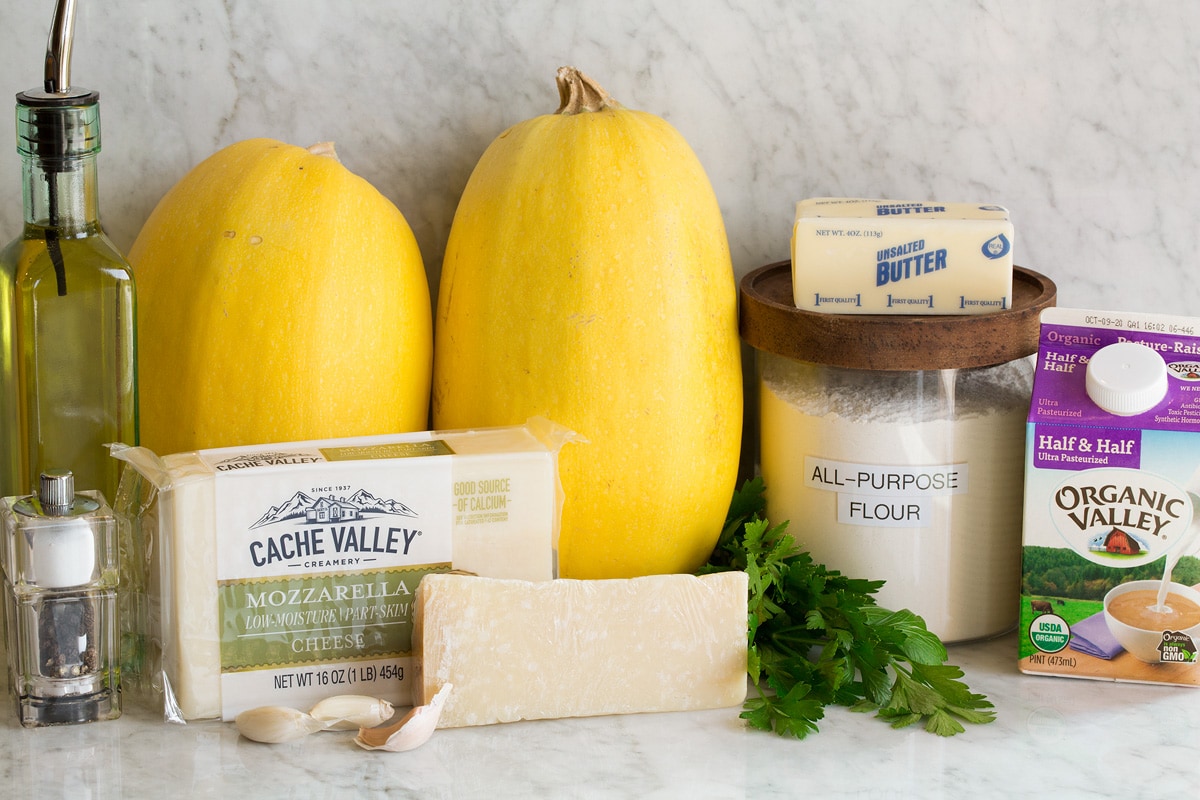 Image of ingredients used to make cheesy spaghetti squash shown. Includes spaghetti squashes, mozzarella, parmesan, half and half, flour, butter, olive oil, garlic, parsley, salt and pepper.