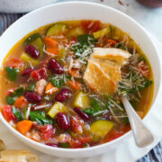 Image of serving of minestrone soup in a white bowl set over white plate. Soup is garnished with parmesan and topped with bread.