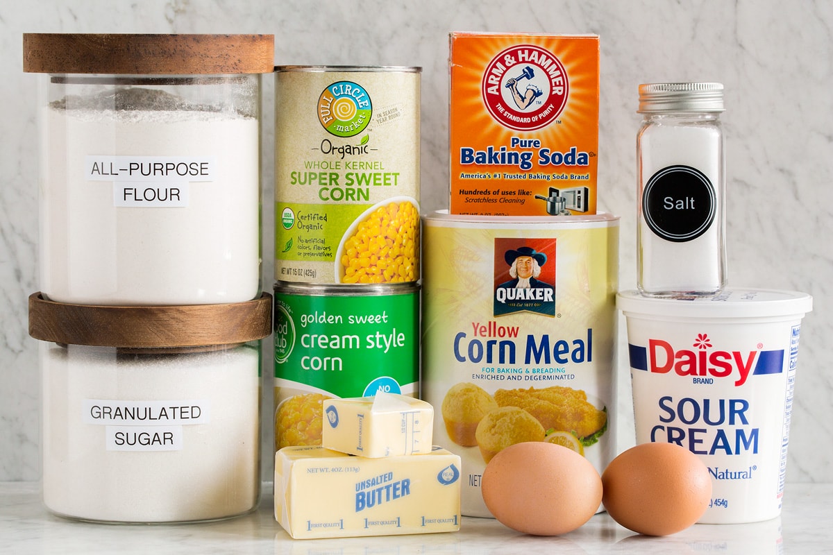 Image of ingredients needed to make corn casserole from scratch. Includes canned corn and creamed corn, flour, sugar, baking soda, sour cream, corn meal, salt, eggs and butter.