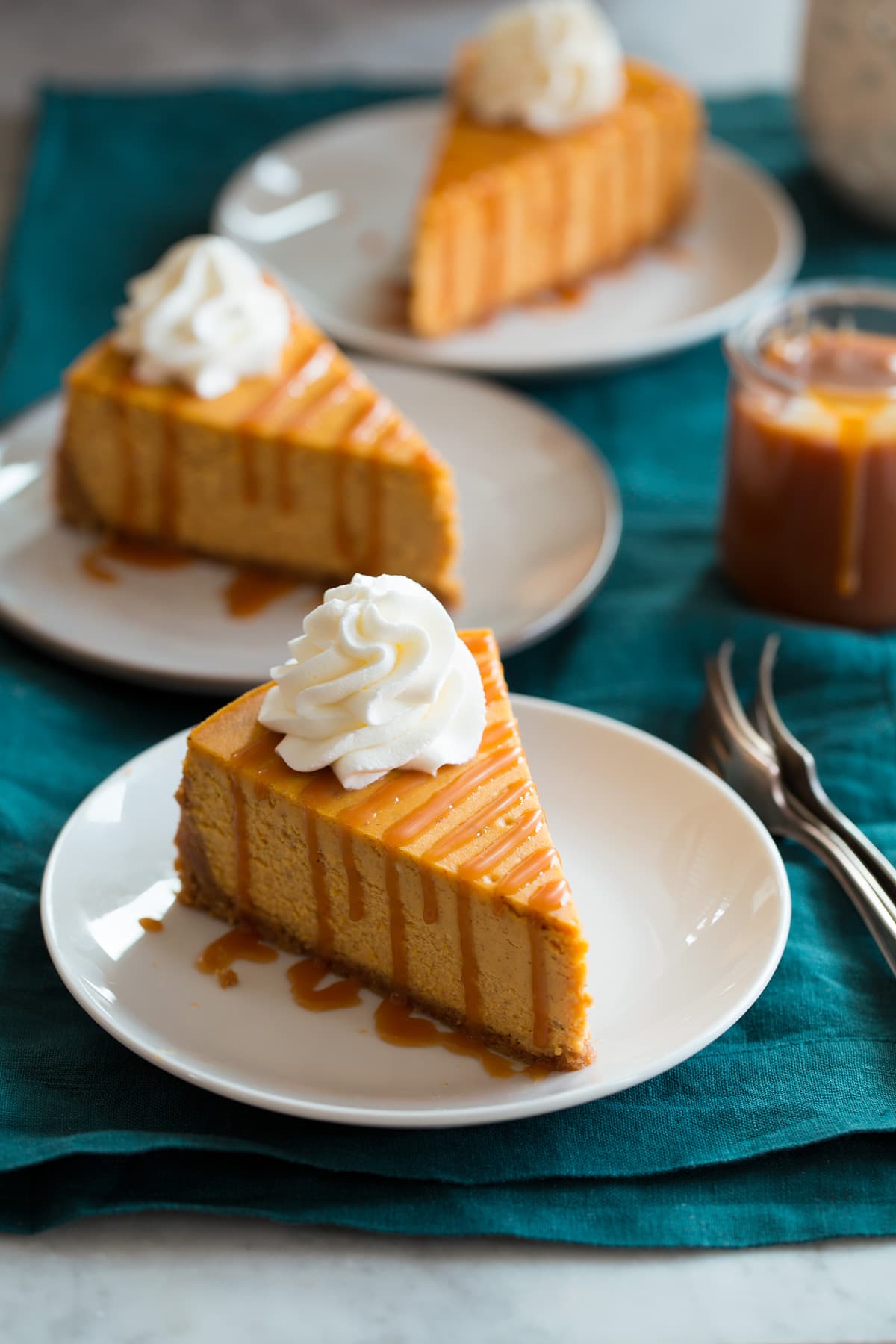 Three slices of pumpkin cheesecake shown on small white dessert plates set over a teal colored cloth.