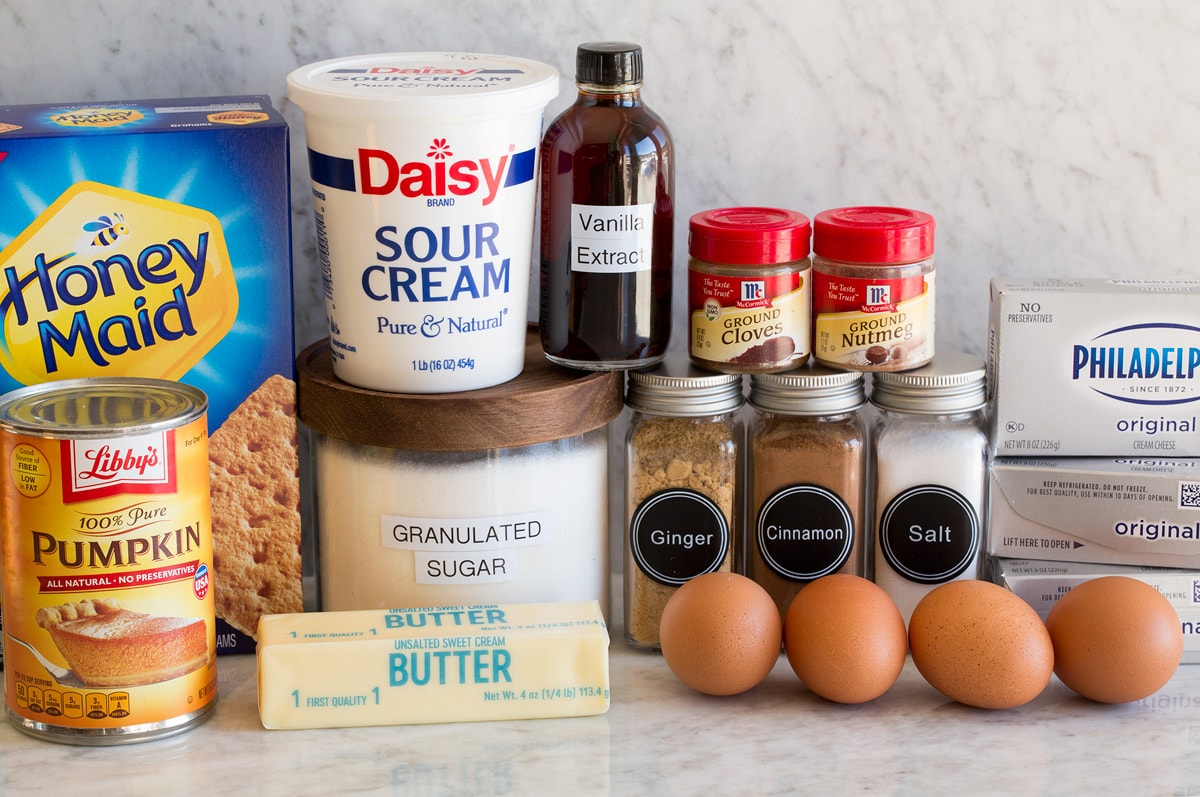 Image of ingredients needed to make pumpkin cheesecake. Includes graham crackers, sugar, butter, cream cheese, eggs, pumpkin, spices, sour cream and vanilla.