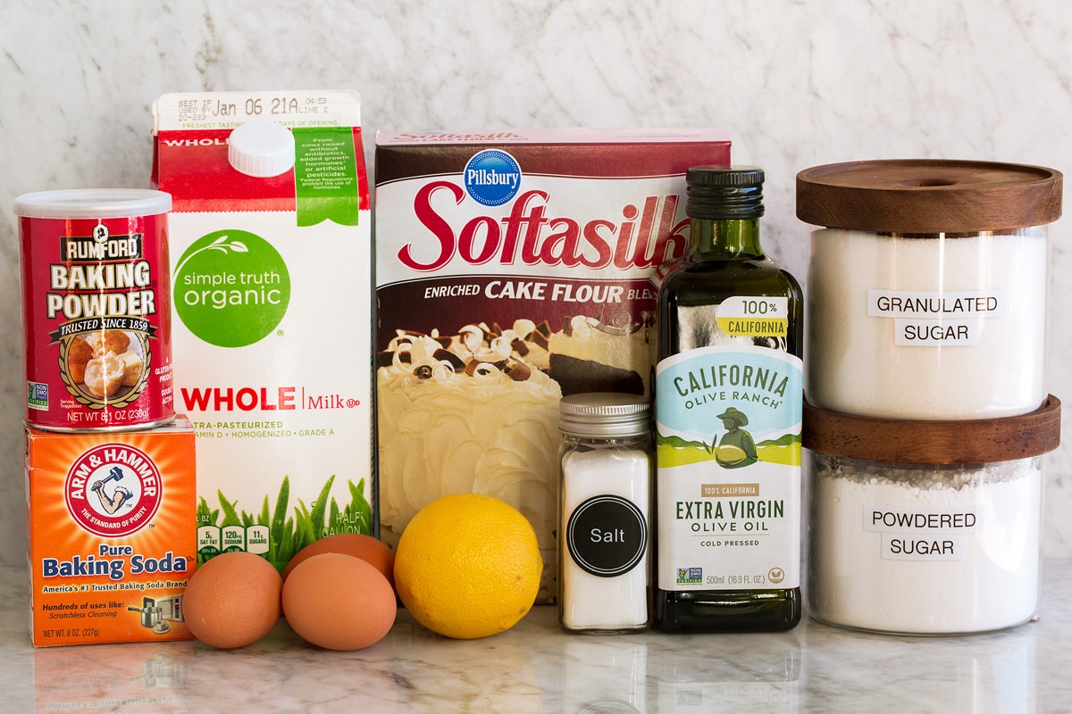 Photo of ingredients used to make olive oil cake. Includes cake flour, baking powder, baking soda, salt, eggs, lemon, extra virgin olive oil, milk, granulated sugar and optional powder sugar.