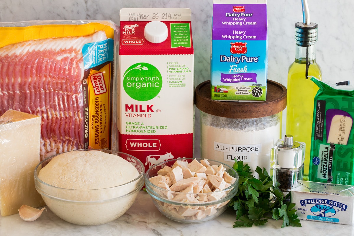 Photo of ingredients for making chicken alfredo pizza. Includes bacon, parmesan, pizza dough, garlic, chicken, parsley, flour, heavy cream, milk, olive oil, mozzarella cheese and butter.