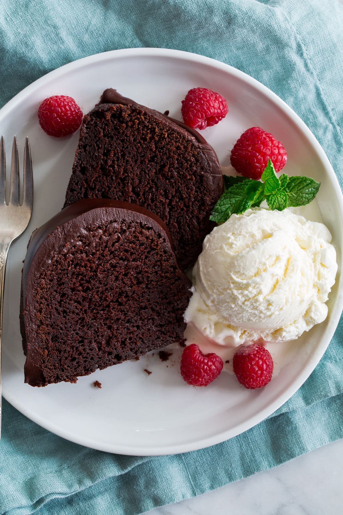 Two slices of chocolate bundt cake on a white plate with a scoop of vanilla ice cream, raspberries and mint to the side.