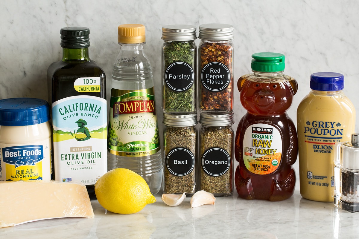 Photo of ingredients used in making homemade Italian dressing. Includes mayonnaise, parmesan, olive oil, lemon, white wine vinegar, parsley, basil, oregano, red pepper flakes, garlic, honey, dijon mustard, salt and pepper. 